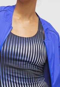 Chaqueta ligera azul sobre una camiseta negra sin mangas con rayas verticales en azul y blanco. Tejido suave con un diseño ajustado.