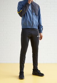 Blau und schwarze Sportjacke mit Reißverschluss vorne, elastischem Taillenbund und schwarzen Jogginghosen mit Logo. Tragen mit schwarzen Sportschuhen.
