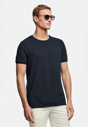 T-shirt en jersey côtelé bleu marine à manches courtes, à encolure ronde, porté avec un pantalon de couleur claire. Modèle portant des lunettes de soleil rondes, fond neutre.