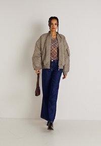 Casaco bomber acolchoado bege, blusa estampada, jeans de perna larga azul-marinho e botas de salto pretas. Modelo leva uma mala em bordeaux profundo.
