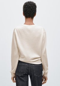 Beige stickad tröja med rund halsringning, ribbade muddar och fåll. Kommer tillsammans med mörka denimjeans, som visar minimalistisk design och textur.