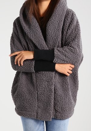 Persona con chaqueta de forro polar sherpa gris de gran tamaño con un amplio cuello y mangas negras superpuestas, combinada con jeans azul claro.