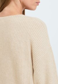 Pull en tricot beige avec une texture côtelée, offrant une encolure large et une coupe décontractée, mettant en valeur des variations subtiles dans l'épaisseur du fil.