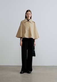 Bež prošiveni trenc kaput s velikim ovratnikom i kopčanjem na gumbe, upareni s crnim širokim hlačama i malom torbicom. Glatka tekstura.