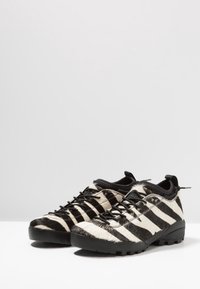 Zapatos a rayas blancas y negras con parte superior texturizada de piel, suela robusta de goma negra y cordones negros. Cuenta con una lengüeta para tirar en el talón.