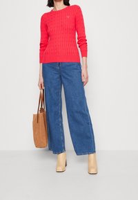 Woman wearing a red cable-knit sweater, blue wide-leg jeans, beige ankle boots, and holding a brown suede tote bag.