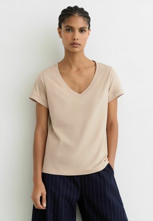 Jonge vrouw met gevlochten haar, gekleed in een beige V-hals top met korte mouwen en marineblauwe broek met krijtstrepen, staand tegen een witte muur.