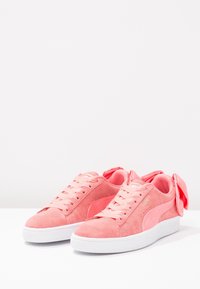 Sneaker in suede rosa con suola in gomma bianca, caratterizzati da un grande fiocco rosa sul tallone e stringhe abbinate. Logo laterale in un rosa più scuro.