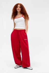 Giovane donna con capelli ricci e rossi, che indossa una canotta bianca, pantaloni rossi larghi e ballerine argentate, in piedi con le mani nelle tasche su sfondo bianco.