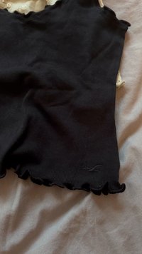 Black ribbed fabric shorts with scalloped edges and a small logo. The texture is soft, and the design is fitted. Background shows a light surface.