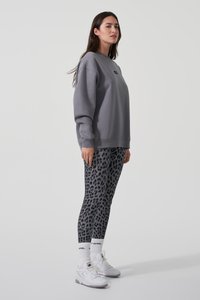 Grauer Sweatshirt mit Rundhalsausschnitt und Logo, kombiniert mit schwarzen Leggings im Leopardendruck und weißen Sneakers. Das Modell steht im Profil.