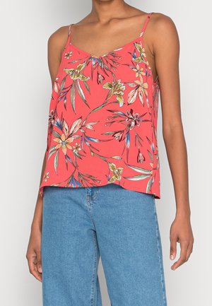 Kobieta ubrana w koralowy top na ramiączkach z żółto-różowym kwiatowym wzorem, w połączeniu z niebieskimi dżinsami, na jednolitym tle.