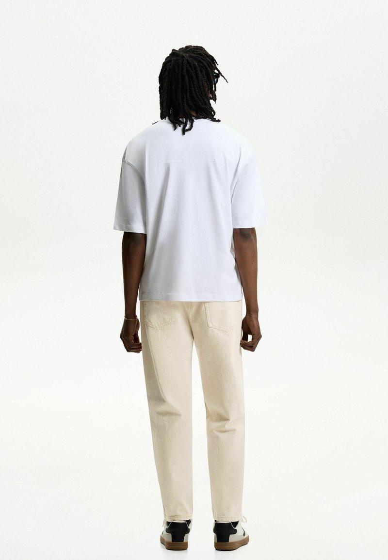 Un homme avec des dreadlocks portant un t-shirt blanc surdimensionné, un pantalon beige et des baskets noir et blanc se tenant de dos sur un fond blanc uni.