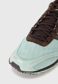 Ljusblå löparsko i mesh med bruna detaljer, texturerad yta och reflekterande inslag. Utrustad med snörning och gummisula.