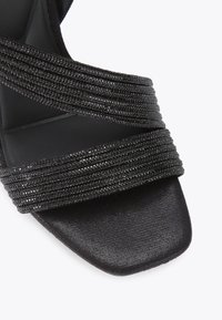 Zwarte hoge hakken sandaal met meerdere gekruiste banden van een textuurstof en een vierkante teenvorm. Gladde voetbed.