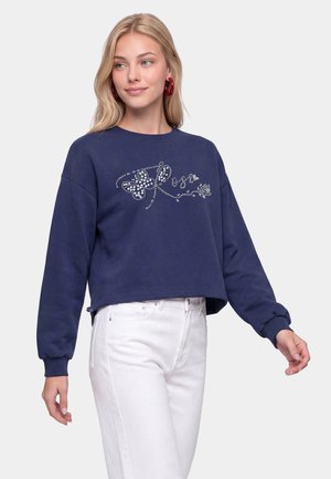 Ung kvinde med blondt hår iført en navyblå sweatshirt med en rhinsten sommerfugl og hvide bukser, der kigger til siden.