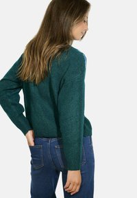 Maglione lavorato a maglia color turchese con un fit rilassato, polsini e orlo a coste. Indossato sopra jeans blu scuro, mostrando un tessuto texture e un design semplice.