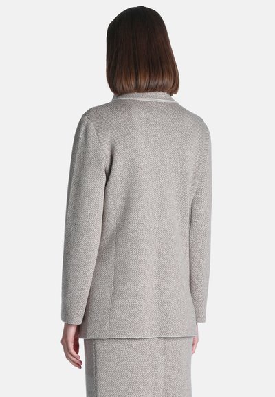 Femme aux cheveux foncés mi-longs porte un blazer gris clair à motif chevrons et une jupe assortie, vue de dos.