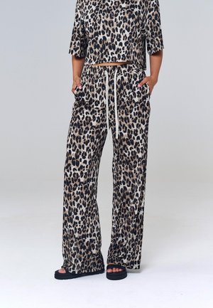 Persoană purtând pantaloni largi cu imprimeu leopard și top asortat cu mâneci medii, stând cu mâinile în buzunare, sandale negre cu platformă vizibile.