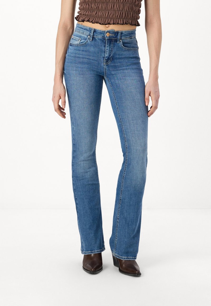Jean flare en denim bleu clair avec un délavage, comportant cinq poches, des passants de ceinture et une fermeture éclair. Portés avec des bottes de cheville marron.