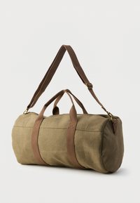 Sac de sport en toile vert olive avec des sangles tissées marron, forme arrondie, fermeture éclair supérieure et texture en tissu. Bandoulière ajustable incluse.