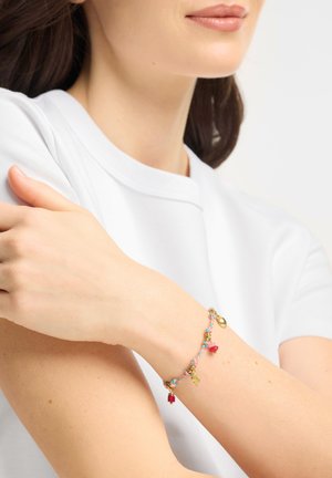 Valkoisessa paidassa oleva nainen, jolla on ranteessaan punottu rannekoru, jossa on kultaisia koristeita ja punaisia helmiä, pitää vastakkaista kättään.