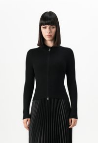 Young woman with straight dark hair wearing a black ribbed zip-up sweater and a black pleated skirt against a plain white background.