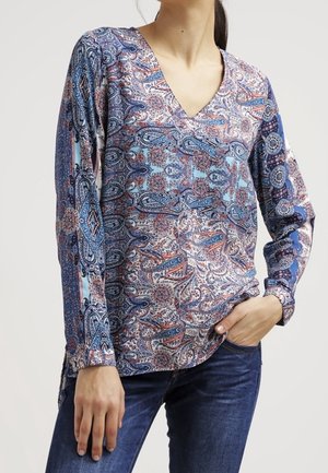 Kvinde iført en langærmet V-hals bluse med et flerfarvet paisleymønster i blå, orange og hvid, kombineret med mørkeblå jeans.