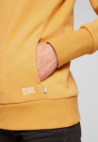Main dans la poche d'un sweat-shirt jaune moutarde avec un bord-côtes et une étiquette blanche Q/S près de la couture de la poche.