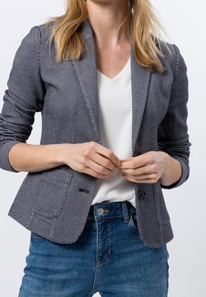 Vrouw draagt een blauw gebloesd colbert, witte blouse en blauwe jeans, staand met gevouwen handen ter hoogte van de taille tegen een effen achtergrond.