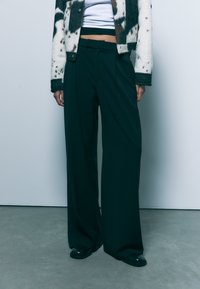 Široke temno zelene hlače iz gladkega materiala, s prostim krojem in subtilnimi pregibi. V kombinaciji z vzorčastim pletenim jopičem.