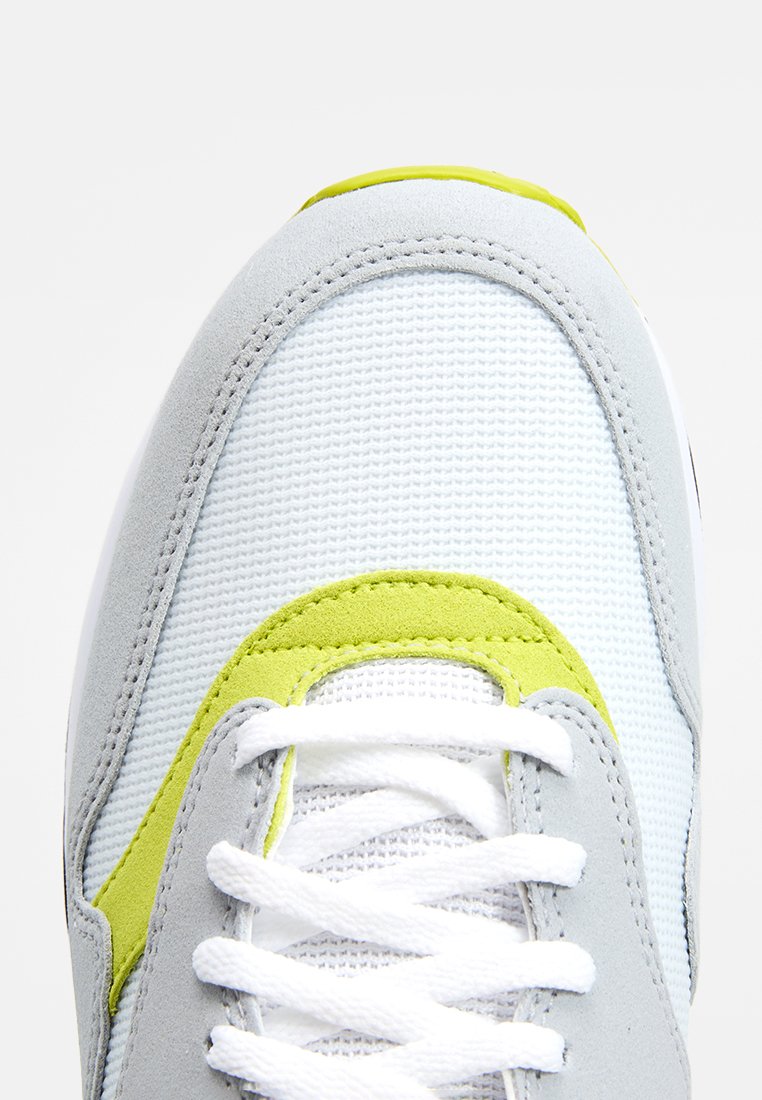 Lichtgrijze sneaker met een witte mesh bovenkant, geel-groene accenten en witte veters. Suède en mesh materialen creëren een textuurrijke uitstraling.