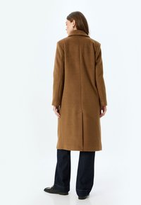 Manteau en laine brun mélangé avec un col structuré, des manches longues et une fente au dos. Associé à un jean noir à jambes larges et des chaussures noires.