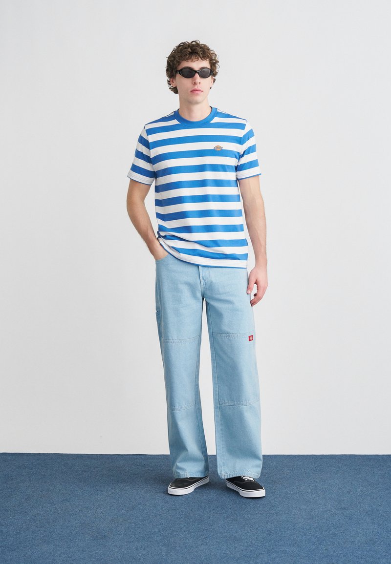 Maglietta a maniche corte a righe bianche e blu, pantaloni a gamba larga azzurri, occhiali da sole neri e sneakers nere. Presenta un piccolo logo sulla maglietta.