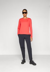 Langermet koralltop med rund hals, kombinert med mørke slim-fit bukser og svarte joggesko, komplett med solbriller.