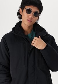 Black padded jacket with a hood, featuring a zip closure and soft texture. Model wears round sunglasses, hand on the jacket’s collar.