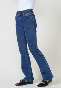 Jeans de denim azul de cintura alta con pierna acampanada. Presenta botones y costuras decorativos, bolsillos frontales y un acabado de textura suave.