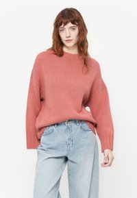 Overdimensjonert strikkegenser i dempet rosa, med ribbestrikkede mansjetter og halslinning, kombinert med høylivede lyseblå denimjeans. Myk tekstur, avslappet passform.