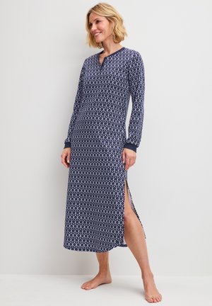 Blonde vrouw die een lange marineblauwe jurk met patroon en lange mouwen en een split aan de zijkant draagt, staat op blote voeten tegen een effen lichte muur.