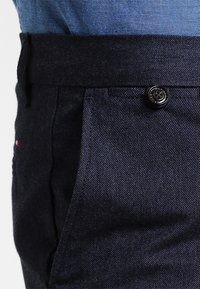 Mörkblå denimbyxor med midja, svart knapp och delvis vy av en instoppad ljusblå skjorta.