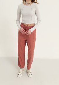 Mujer con una blusa corta de manga larga blanca, pantalones de tiro ajustable en color óxido, zapatillas blancas y un collar choker plateado, de pie frente a un fondo liso.