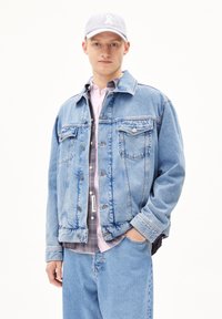 Lichtblauwe spijkerjack met twee borstzakken, zilveren knopen en een relaxte fit, gelaagd over een grijze blouse en een lichtroze shirt.