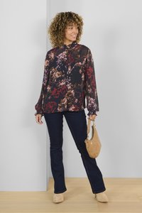 Blusa nera fluida e trasparente con stampa floreale in borgogna, rosa e verde; maniche lunghe con polsini a volants. Abbinata a jeans scuri e borsa chiara color beige.
