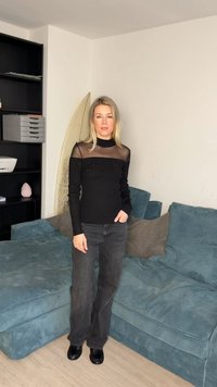 Femeie stând într-un living, purtând un top negru transparent, blugi închiși la culoare și pantofi negri, lângă o canapea de catifea teal și o placă de surf.