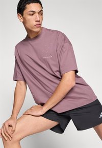 Μπλουζάκι crewneck από βαμβάκι σε ανοιχτό μωβ χρώμα, με μικρό λευκό γράφημα στο μπροστινό μέρος που γράφει "τρέξε με το μυαλό". Συνδυάζεται με μαύρο σορτς.