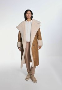 Długi brązowy płaszcz ze shearlingu z puszystym kremowym kołnierzem i mankietami. Nosi się go na jasnym hoodies i beżowych spodniach, w połączeniu z jasnobrązowymi butami na sznurówki.