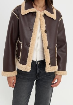 Kvinde iført en mørkebrun jakke i imiteret læder med beige shearlingkant, knappet foran, hvid top og mørkegrå jeans.