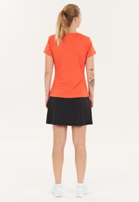 Kvinde står med ryggen til iført en orange kortærmet atletisk t-shirt, sort nederdel, hvide sneakers og hestehale frisyr, med synlige arm tatoveringer.