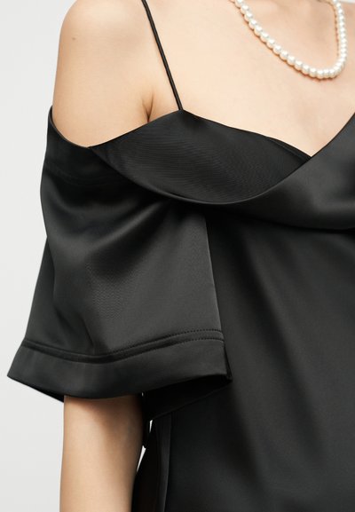 Γυναίκα που φοράει μαύρο σατέν φόρεμα με ακάλυπτους ώμους και λεπτές τιράντες, και περιδέραιο με πέρλες, μπροστά σε απλό φόντο.