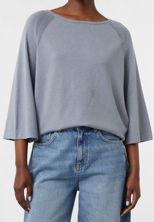 Løs, lysegrå sweater med vide ærmer og tekstureret skulderdetalje, båret med højtaljede lyseblå jeans.